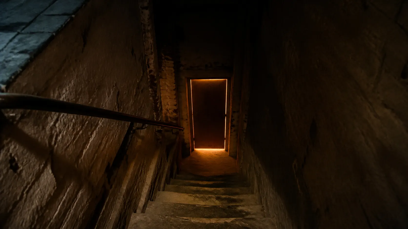 A narrow basement stairwell of painted brick, worn treads descending to a plain unmarked door with warm light leaking around its edges.